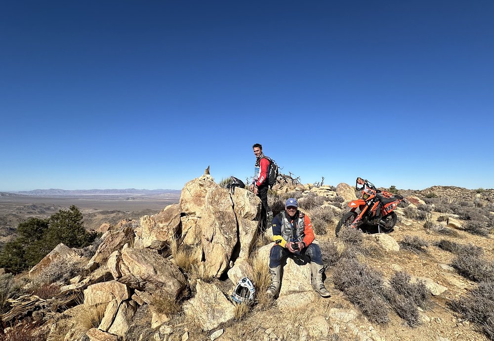 Ari and a friend with their dirt bikes on top of the rocky peak of a mountain in the desert under a clear blue sky