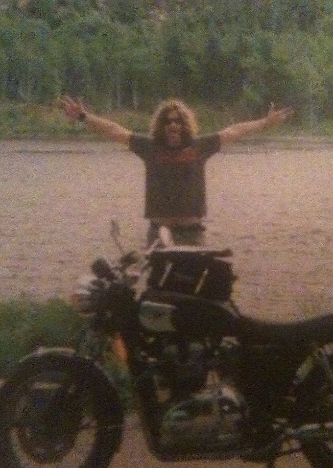 old grainy photo of young Spurgeon with long hair raising his arms and standing behind his Bonneville and in front of a lake