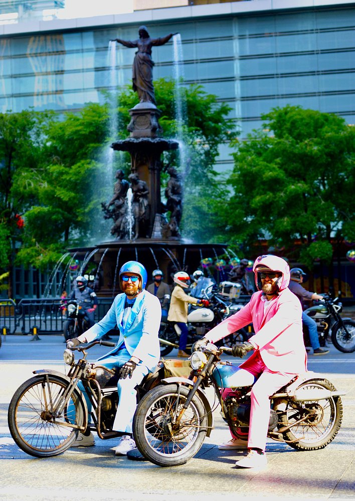riders in light blue and pink suits on motorcycles in front of a fountain in the park