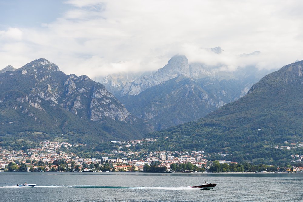 a speed boat crosses Lake Como with the town on the opposite shore and mountains in the background