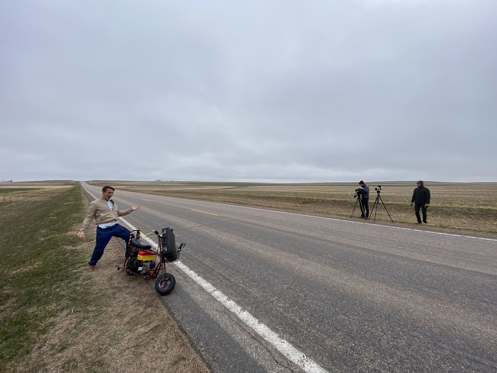 Man standing next to a mini bike on the side of a straight road.