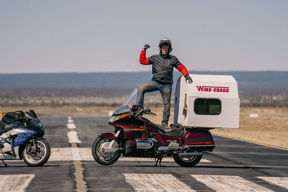 A man standing on the seat of a 1989 Honda Gold Wing with a small house on the back, aka the Wing-ebago.