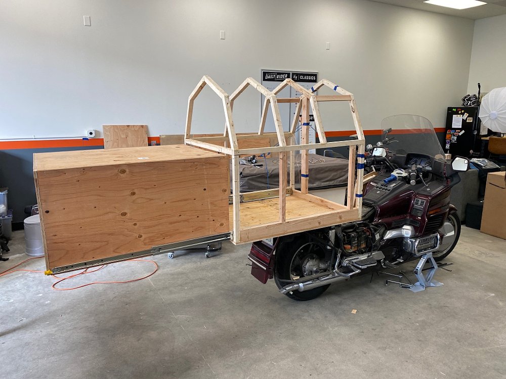 A 1989 Honda Gold Wing with framing for a small building attached to the back, and a drawer-like box extending out the back of the wood-framed building.