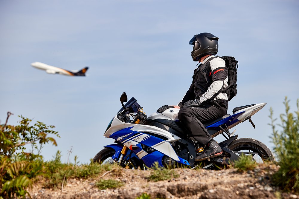A photo of a rider on a sportbike parked, watching an airline jet fly overhead
