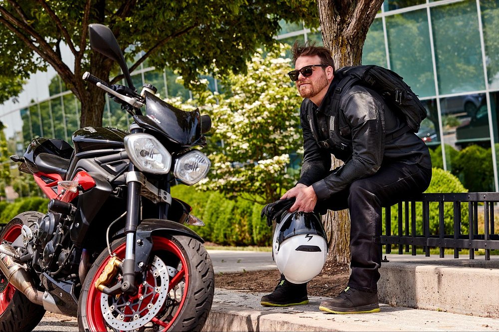 A motorcycle rider sits in the shade of a tree on a city street, bike in foreground