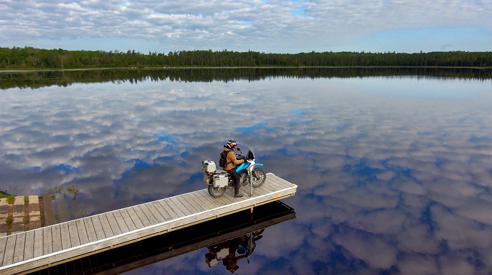 rider on a motorcycle on the end of a dock in a lake with clouds reflected in the water