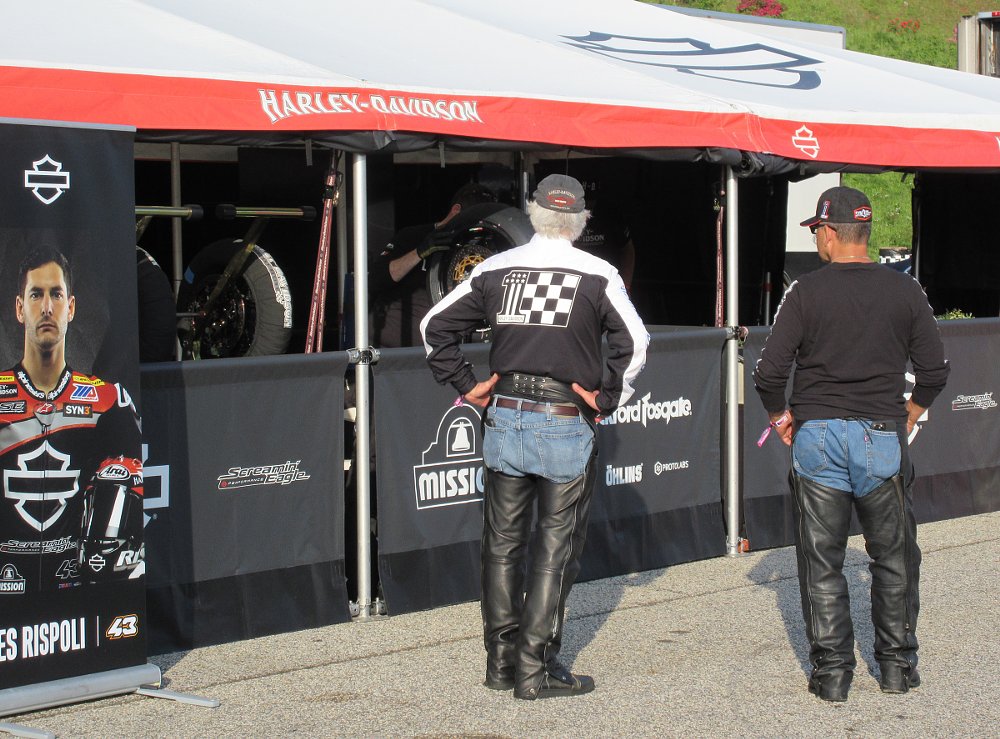 two men wearing Harley-Davidson-branded clothes watching mechanics inside the Harley race team tent
