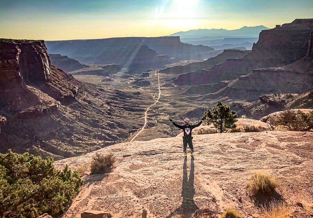 photo of rider in off-road motorcycle gear standing on a rock with a view of a trail and desert cliffs and mountains in the background
