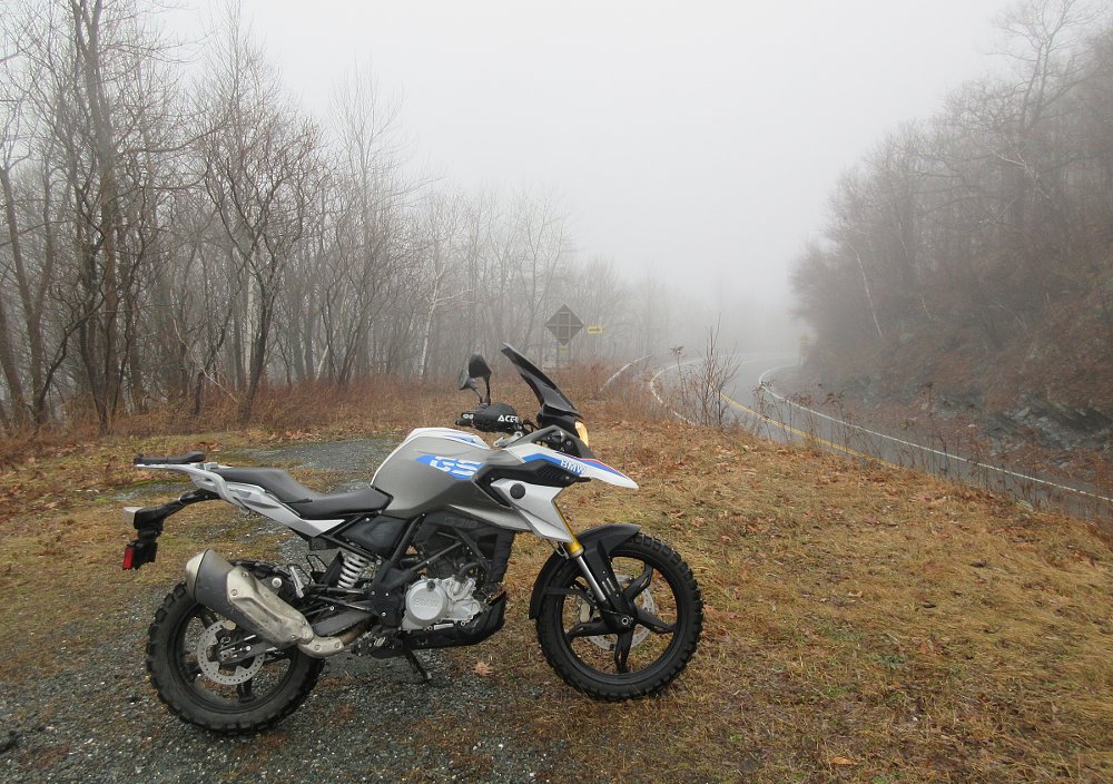 BMW G 310 GS parked atop a mountain with a curving road in the background and low clouds and fog