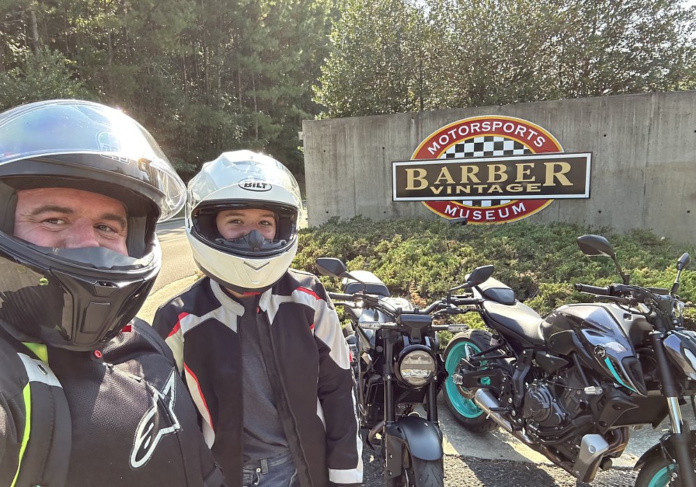 father and daughter still wearing their helmets in a selfie in front of the Barber Vintage Motorsports Museum sign