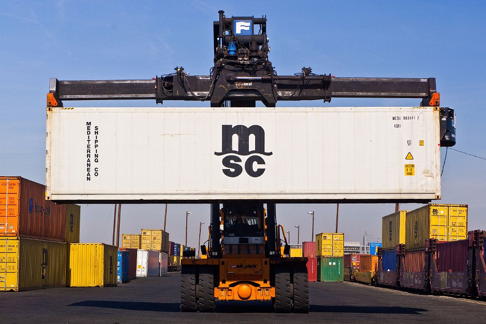A fork lift carries a shipping container through a container yard.